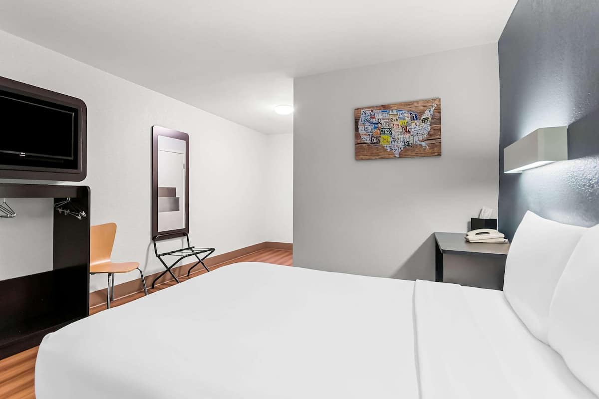A hotel room with a bed, a chair, a desk with a phone, a mirror, and a wall-mounted TV.