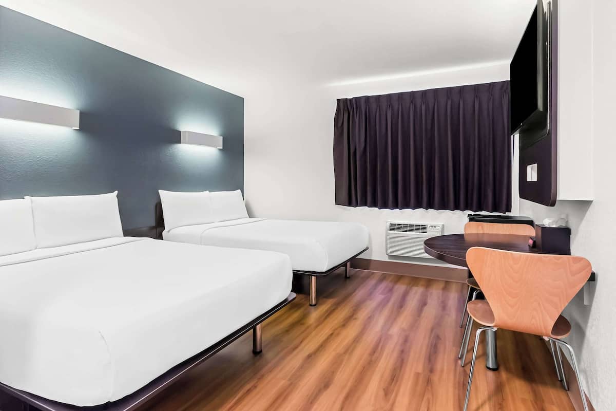 A hotel room with two beds, a desk, a chair, a flat-screen TV, and a window with curtains.