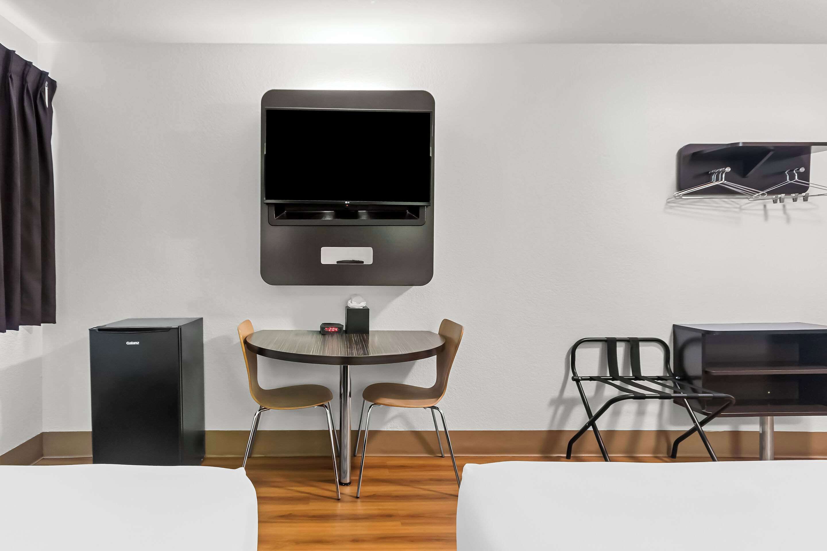 Habitación de hotel pequeña y funcional con cama, escritorio, silla, mesita y televisor montado en la pared.