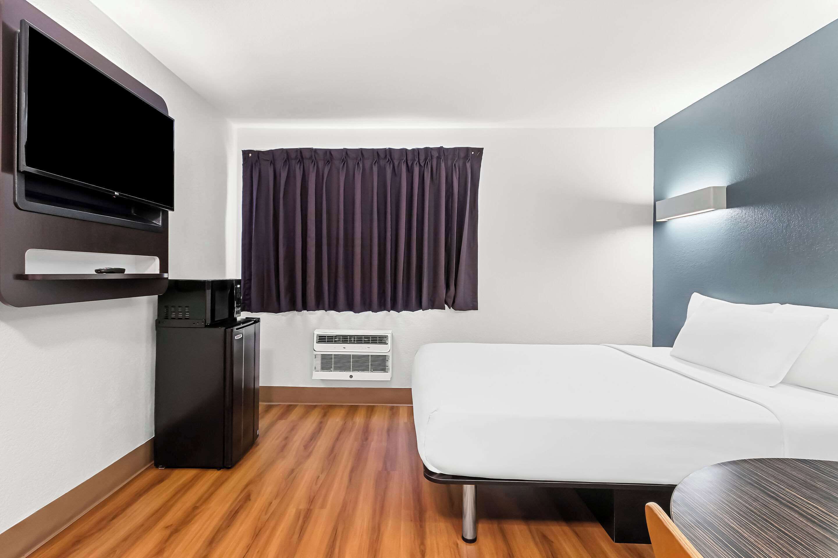 A hotel room with a bed, a TV, a mini-fridge, a heater, and a window with curtains.