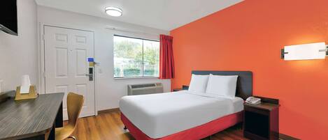 A hotel room with a bed, a desk, a chair, a nightstand, and a window with a curtain.