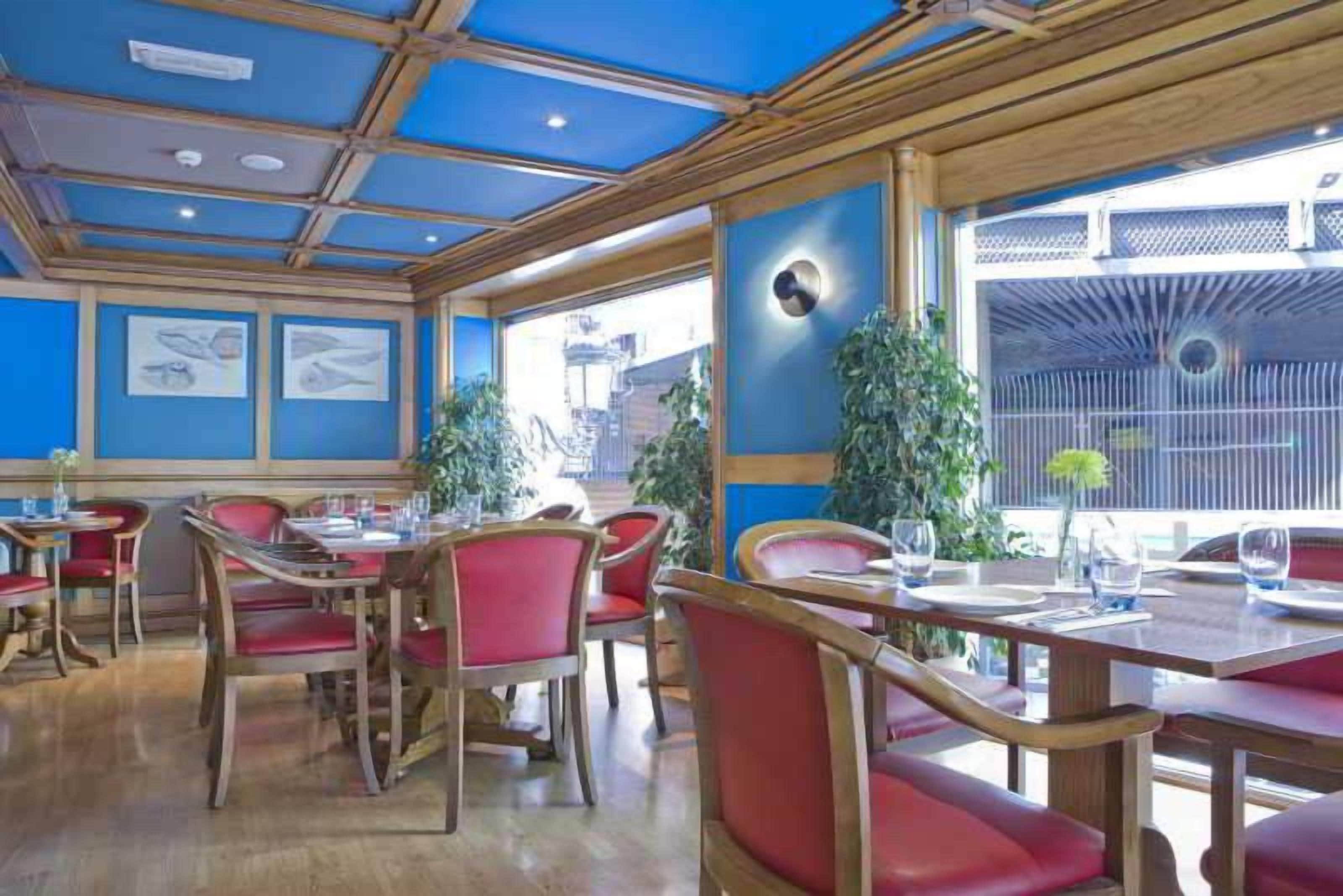 Un restaurante con mesas de madera, sillas rojas, ventanas amplias y paredes azules.