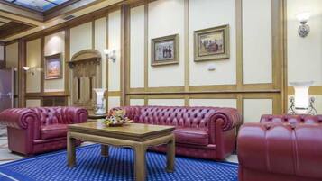 A hotel lobby with a blue patterned carpet, red leather sofas, a wooden coffee table, and framed artwork on the walls.