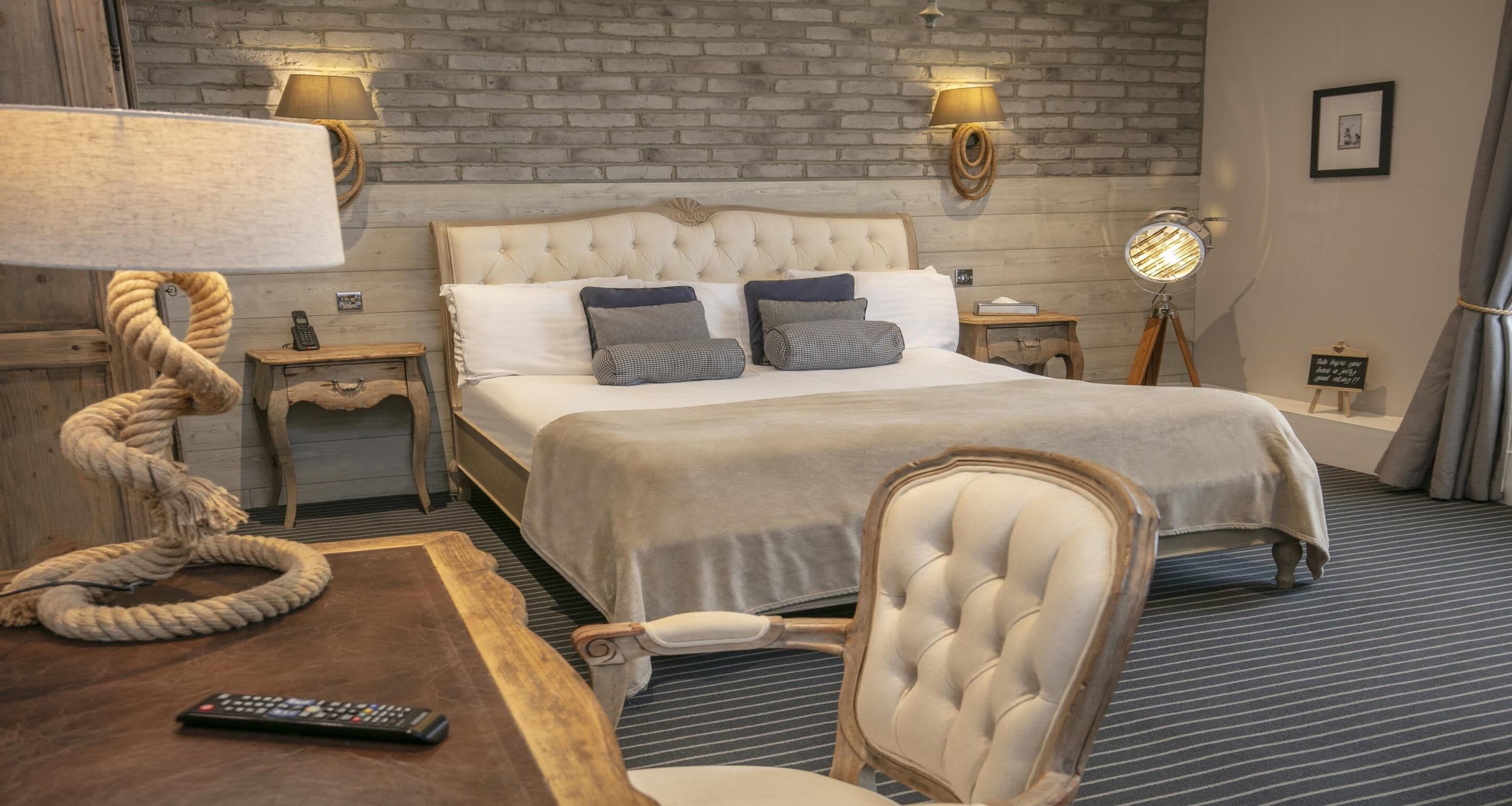 A hotel room with a large bed, a wooden side table, a chair, a lamp, a bedside table, and a wall-mounted lamp.