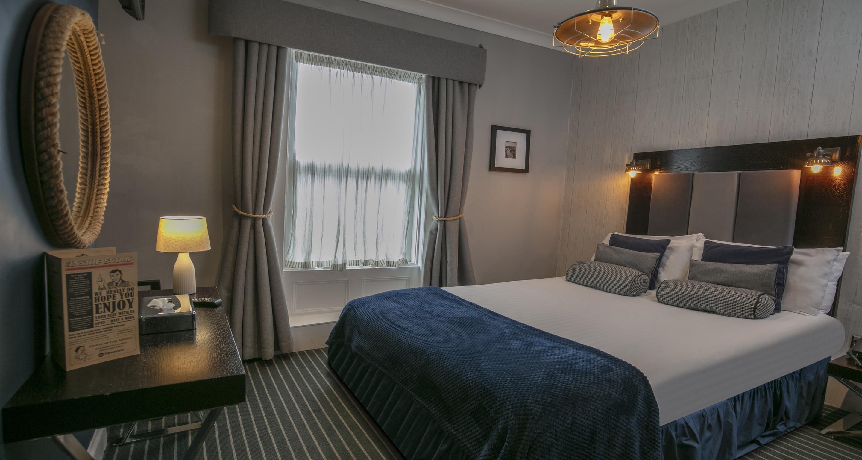 Una habitación de hotel con una cama grande, un escritorio con lámpara, una ventana con cortinas y un cabecero con luces.
