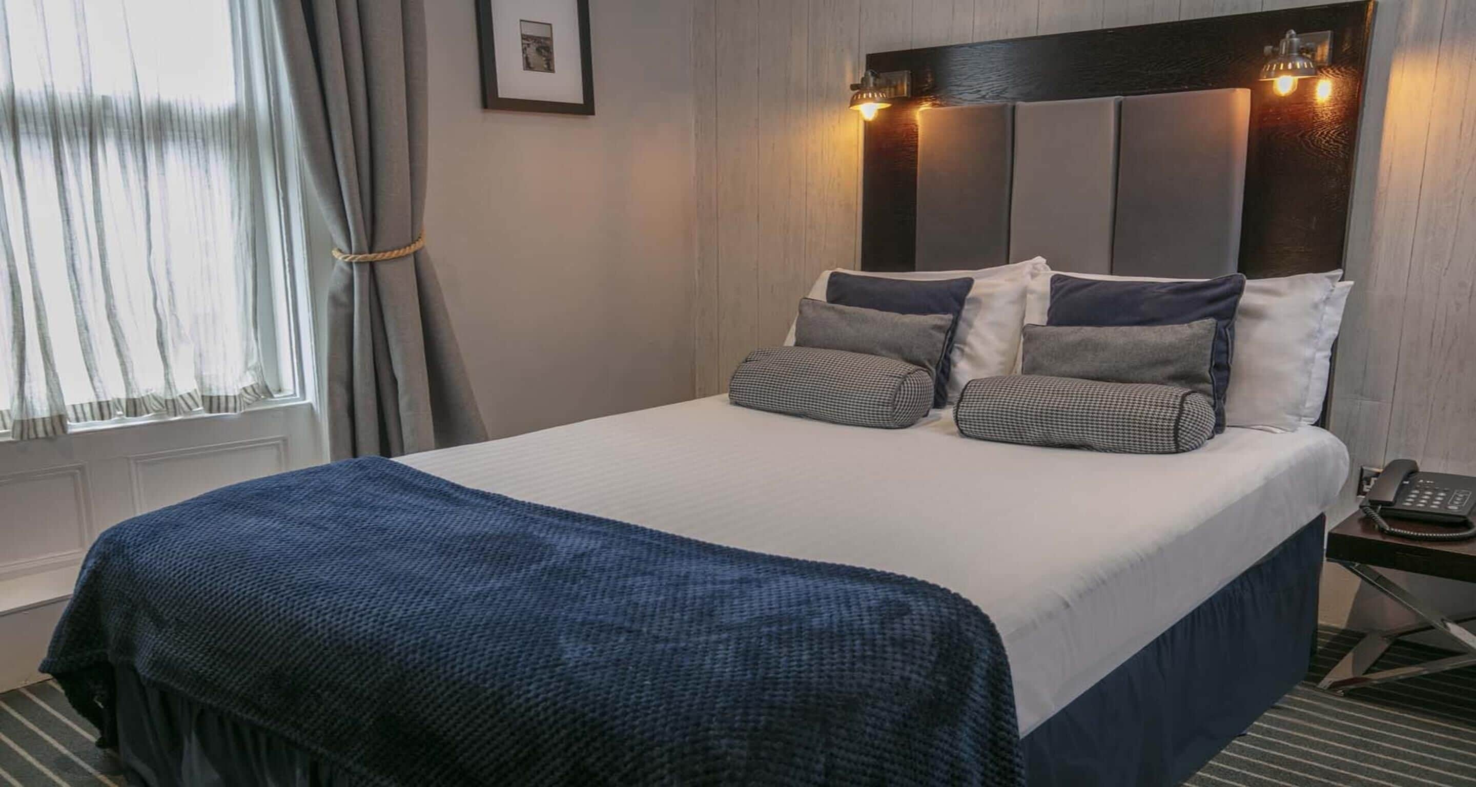 A neatly made bed with a blue blanket, a headboard with lights, and a framed picture on the wall.