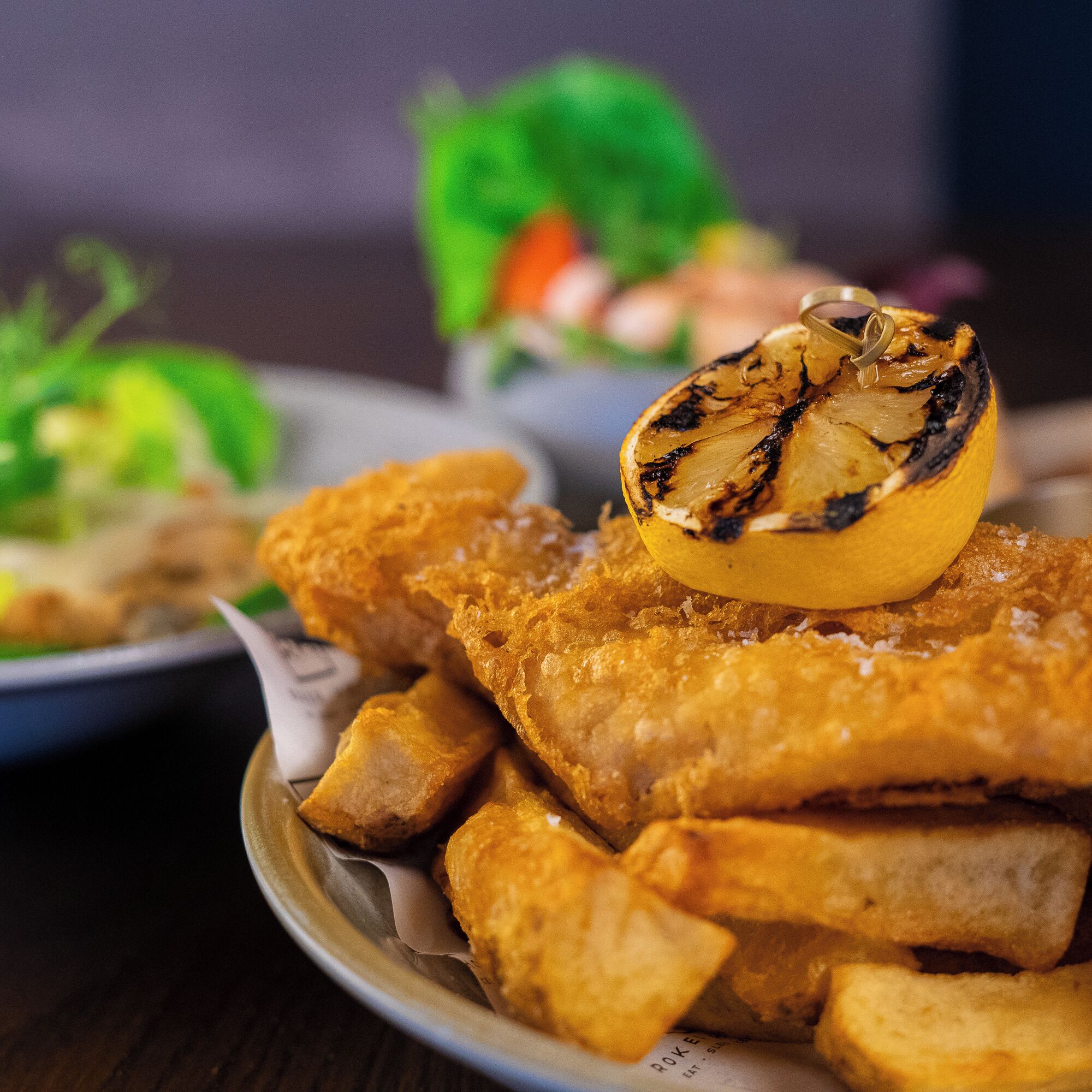 A plate of fried fish fillets with a grilled lemon slice on top, accompanied by golden-brown potato wedges.