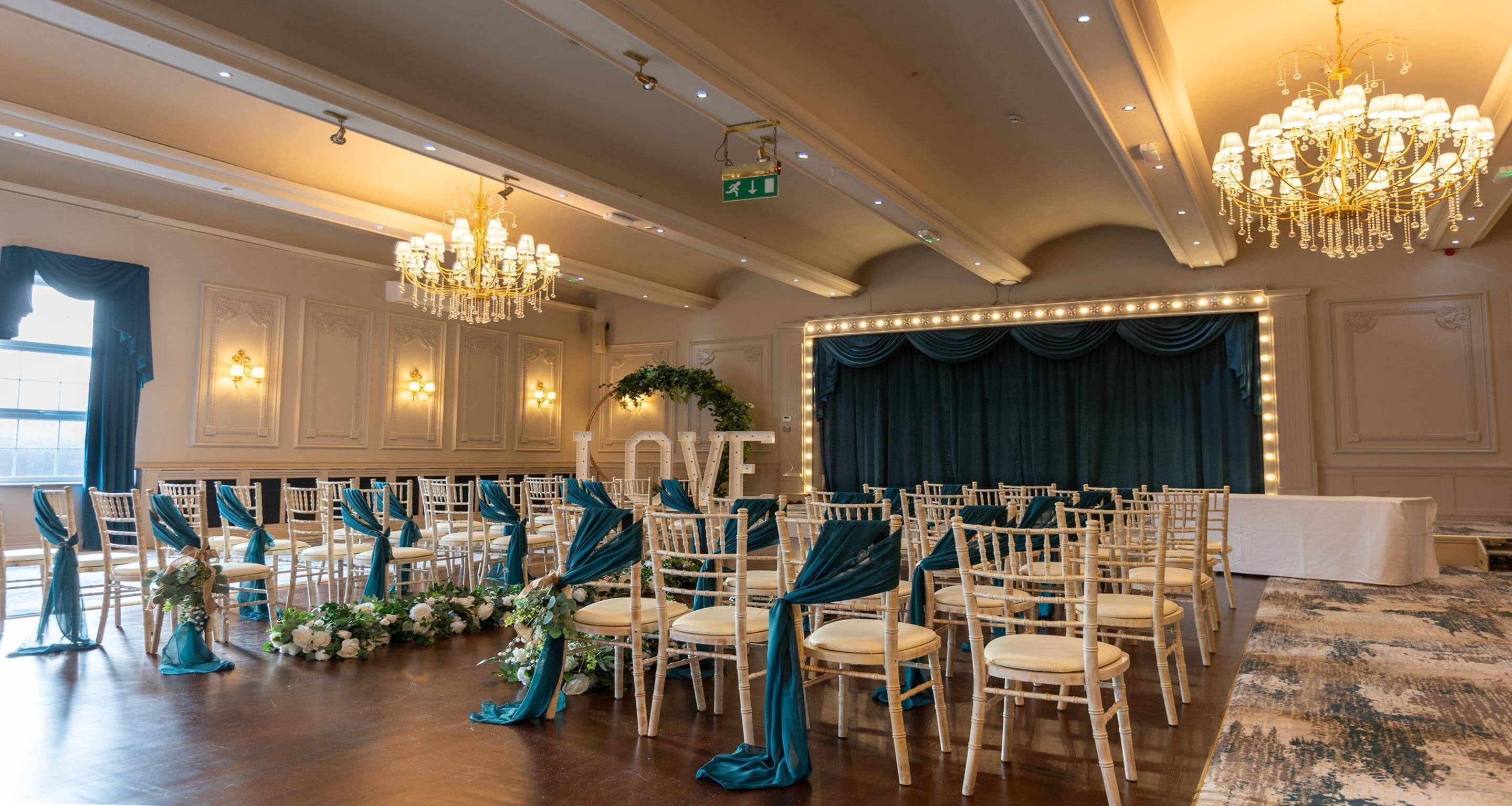 A banquet hall with rows of white chairs, teal drapes, and chandeliers.