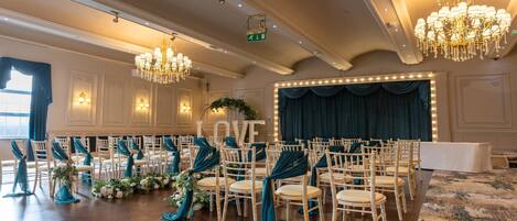 A banquet hall with rows of white chairs, teal drapes, and chandeliers.