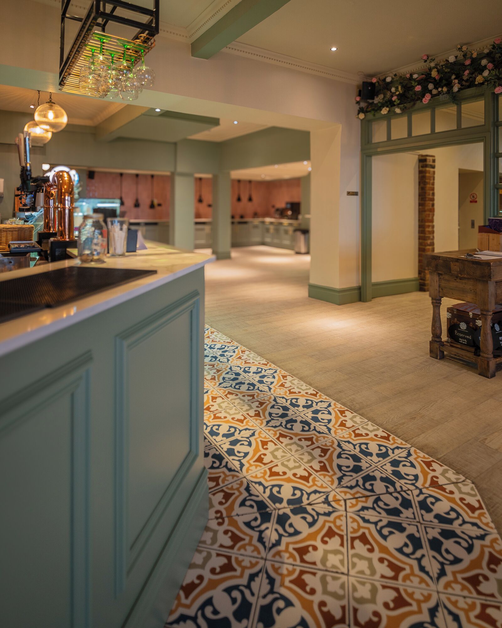 Un hall de hotel con suelo de baldosas estampadas, una zona de bar con detalles de cobre y un arreglo floral decorativo en la pared.