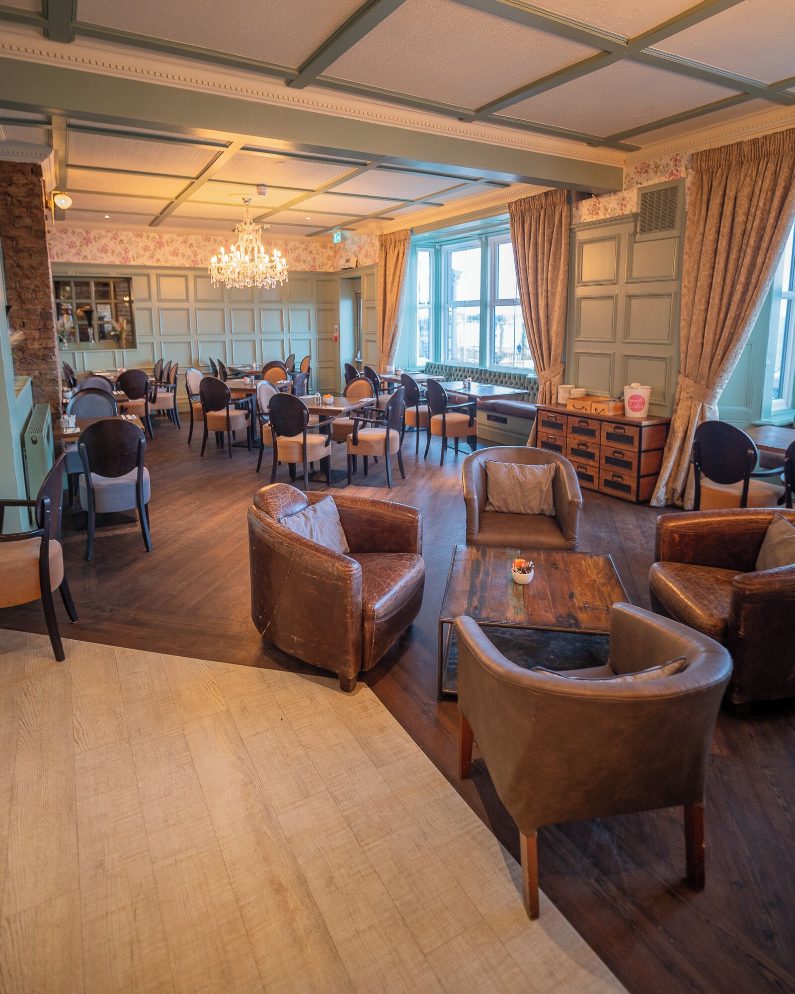 A spacious lobby with leather armchairs, wooden tables, and a chandelier.