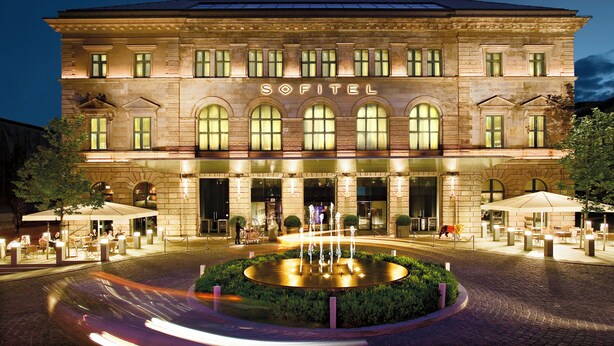 A Sofitel hotel with a fountain in the courtyard, outdoor seating, and a well-lit entrance. - Sofitel Munich Bayerpost (Munich)