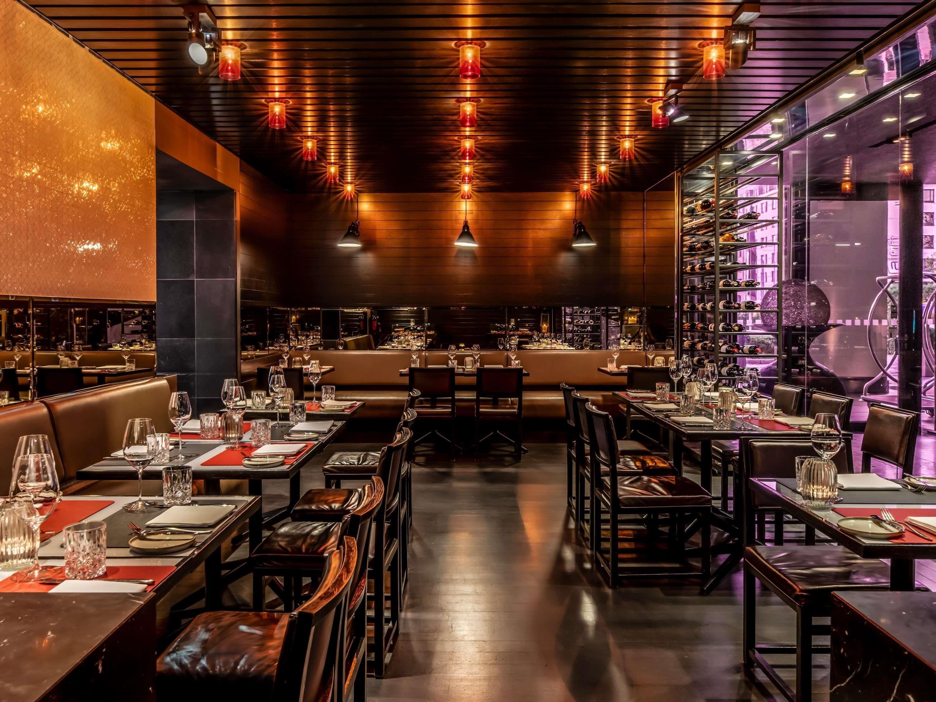 Un restaurante con varias mesas y cabinas, iluminación cálida y una estantería de vinos visible a través del ventanal.