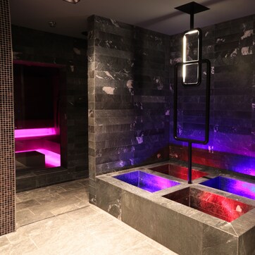 A modern bathroom with a central heated stone bath, a glass enclosure, and a black metal structure above.