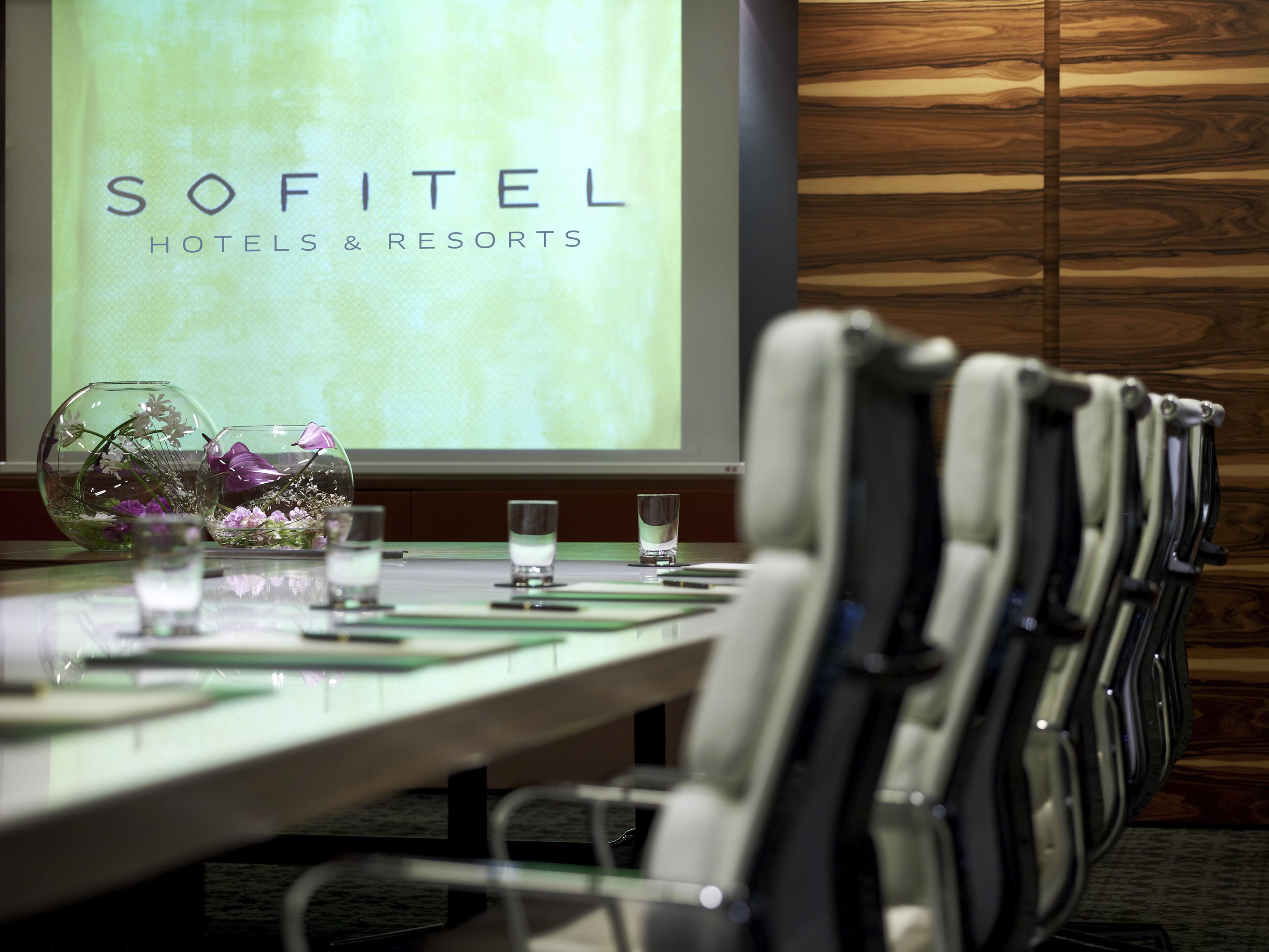 Ein Konferenzraum mit einem langen Tisch, Stühlen, einer Projektionsleinwand mit dem Schriftzug "SOFITEL HOTELS & RESORTS" und dekorativen Glasvasen mit Blumen.