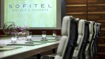 Ein Konferenzraum mit einem langen Tisch, Stühlen, einer Projektionsleinwand mit dem Schriftzug "SOFITEL HOTELS & RESORTS" und dekorativen Glasvasen mit Blumen.