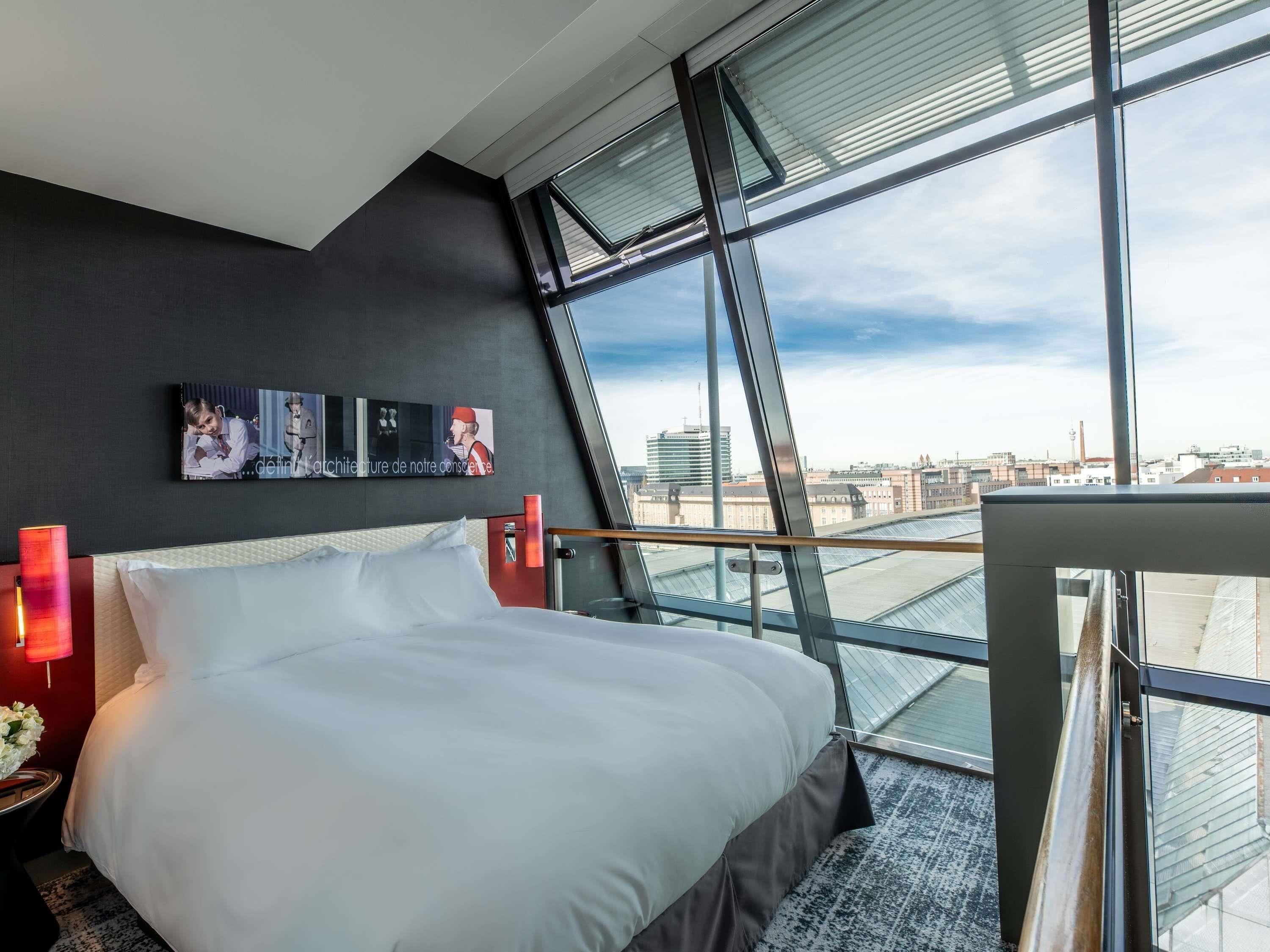 A modern hotel room with a large bed, a city view through floor-to-ceiling windows, and a wall-mounted artwork.