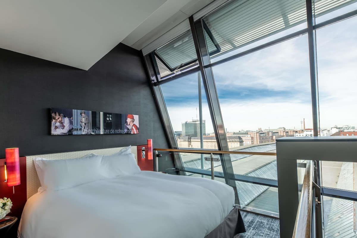 A modern hotel room with a large bed, a city view through floor-to-ceiling windows, and a wall-mounted artwork.