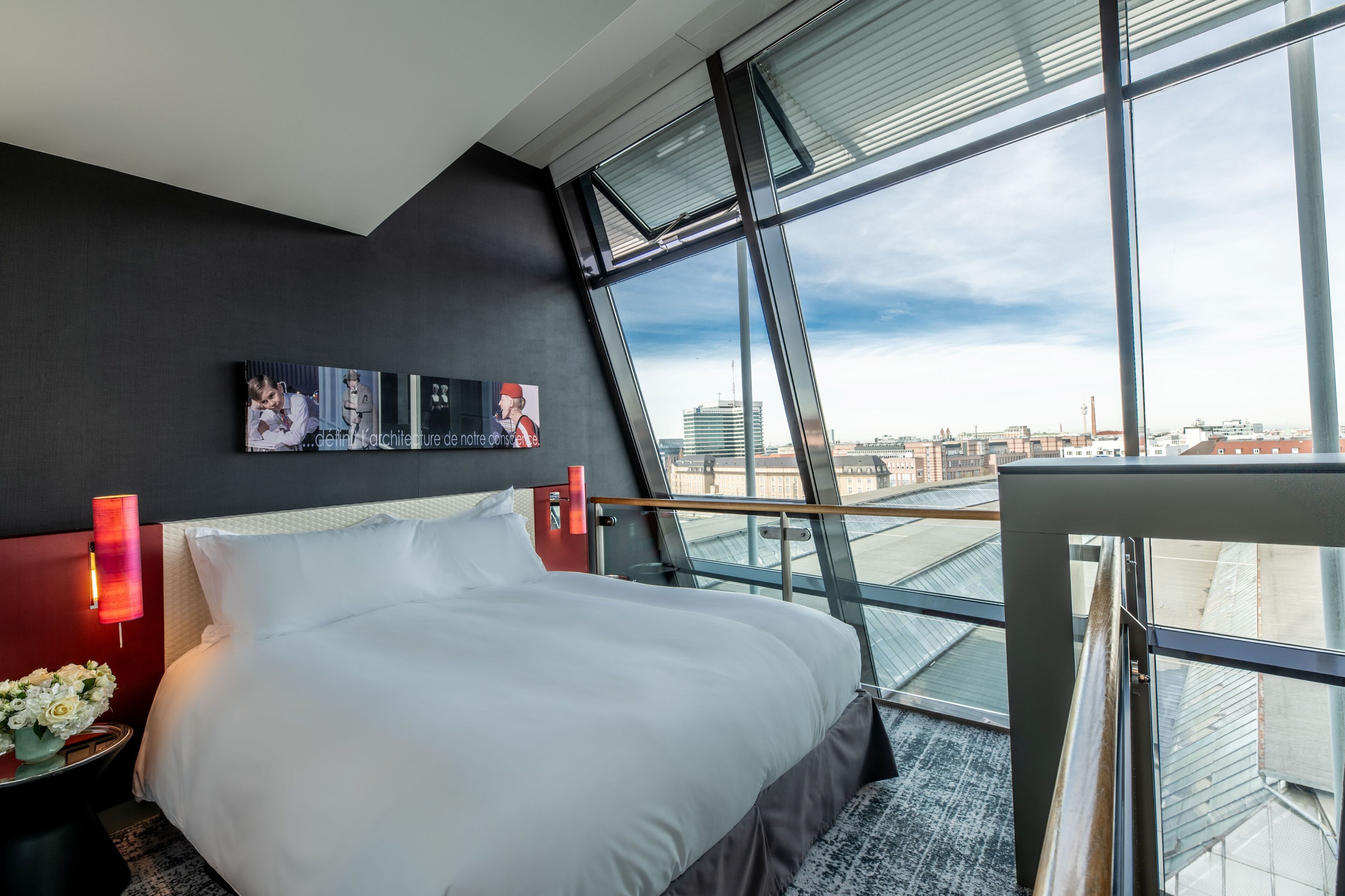A modern hotel room with a large bed, a nightstand, a red lamp, and a city view through large windows.