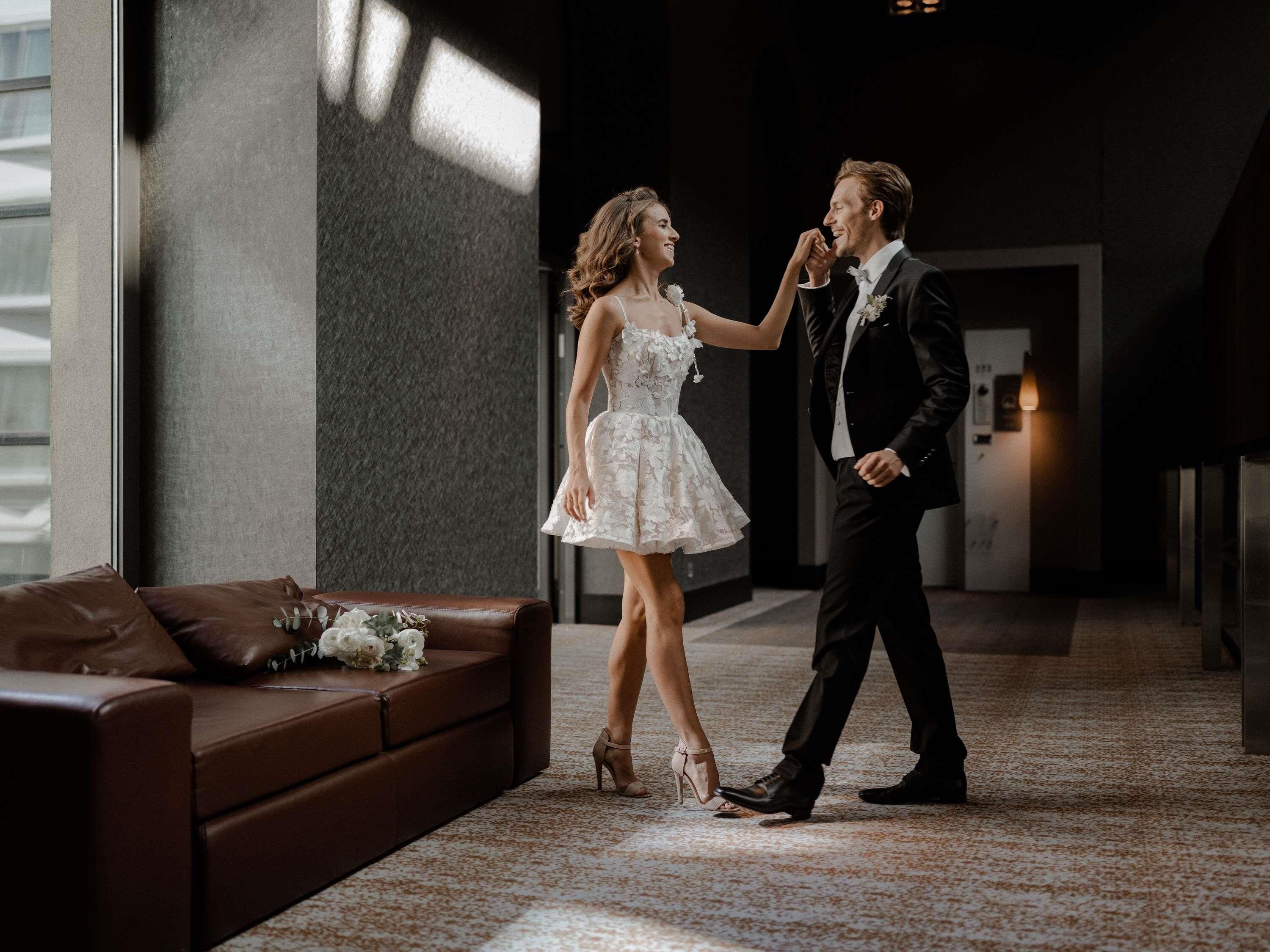 A couple dancing in a modern hotel lobby with a brown leather couch and a bouquet of flowers.