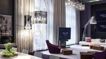 Ein modernes Hotelzimmer mit einem großen Flachbildfernseher, einer Couch, einem Esstisch mit Stühlen und einem dekorativen Kronleuchter.