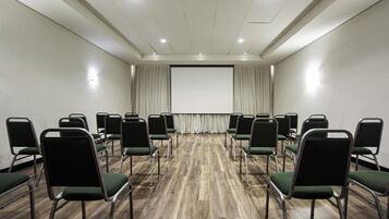 Un salón de conferencias con filas de sillas, una pantalla de proyector y piso de madera.