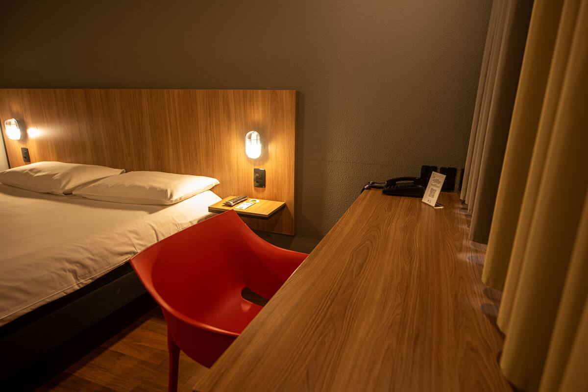 Ein Hotelzimmer mit einem hölzernen Kopfteil, einem Bett mit weißen Leinentüchern, einem roten Sessel und einem Holztisch mit Telefon und Notizblock.