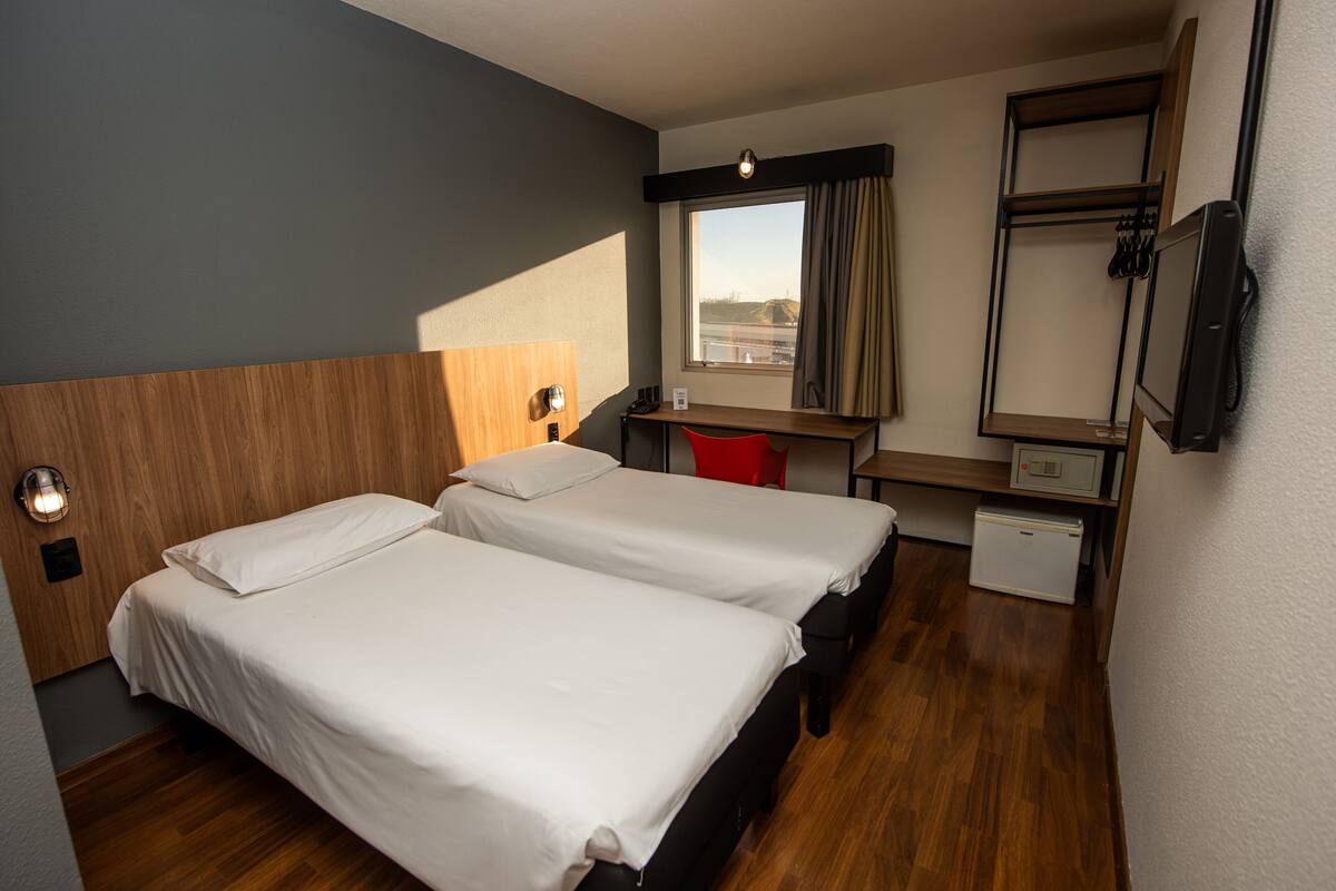 Ein Hotelzimmer mit zwei Betten, einem Schreibtisch, einem Stuhl, einem kleinen Kühlschrank und einem Fenster mit Vorhängen.