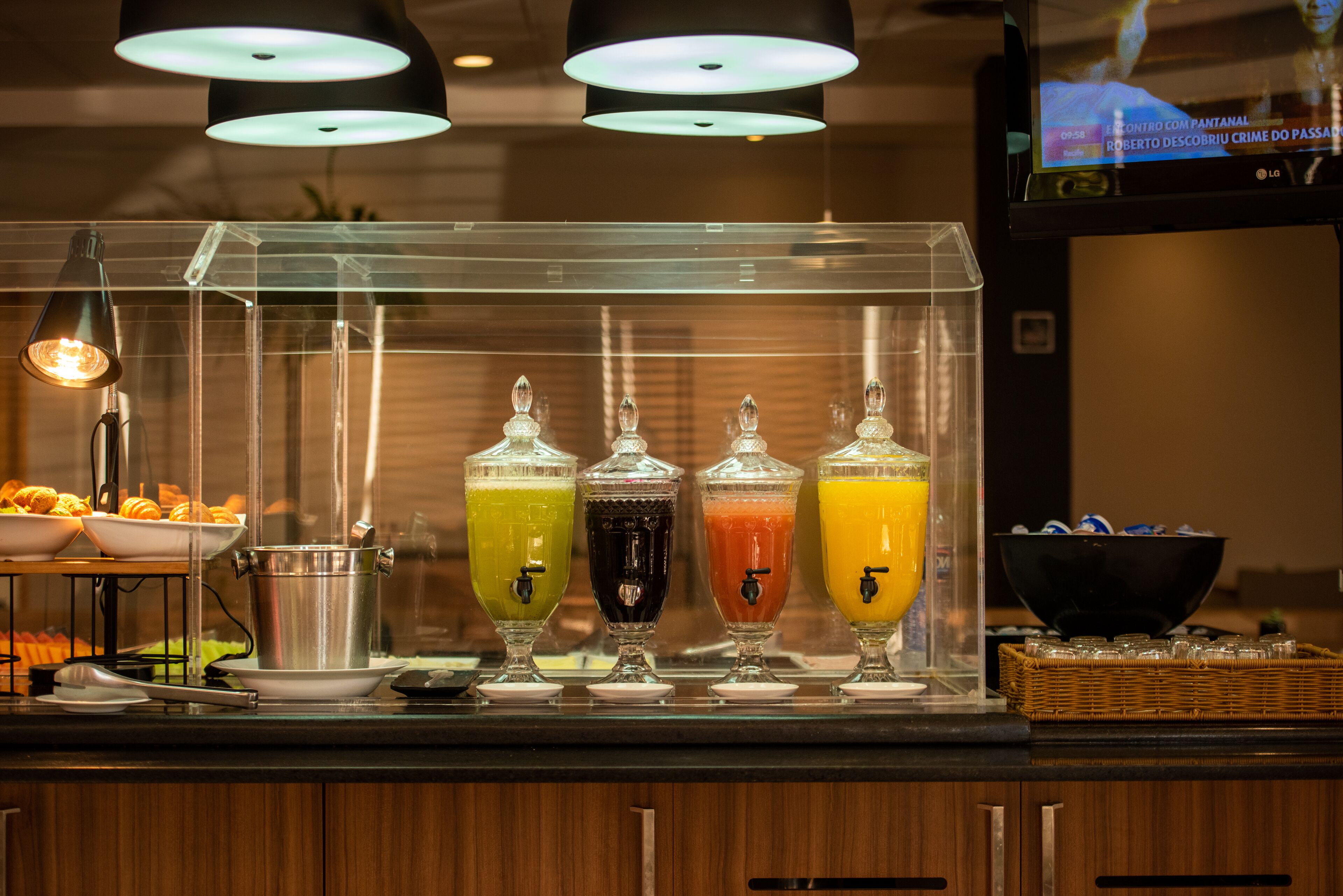 Un bar de zumos con cuatro dispensadores de bebidas de colores distintos, una cesta de pan y una pantalla de televisión al fondo.