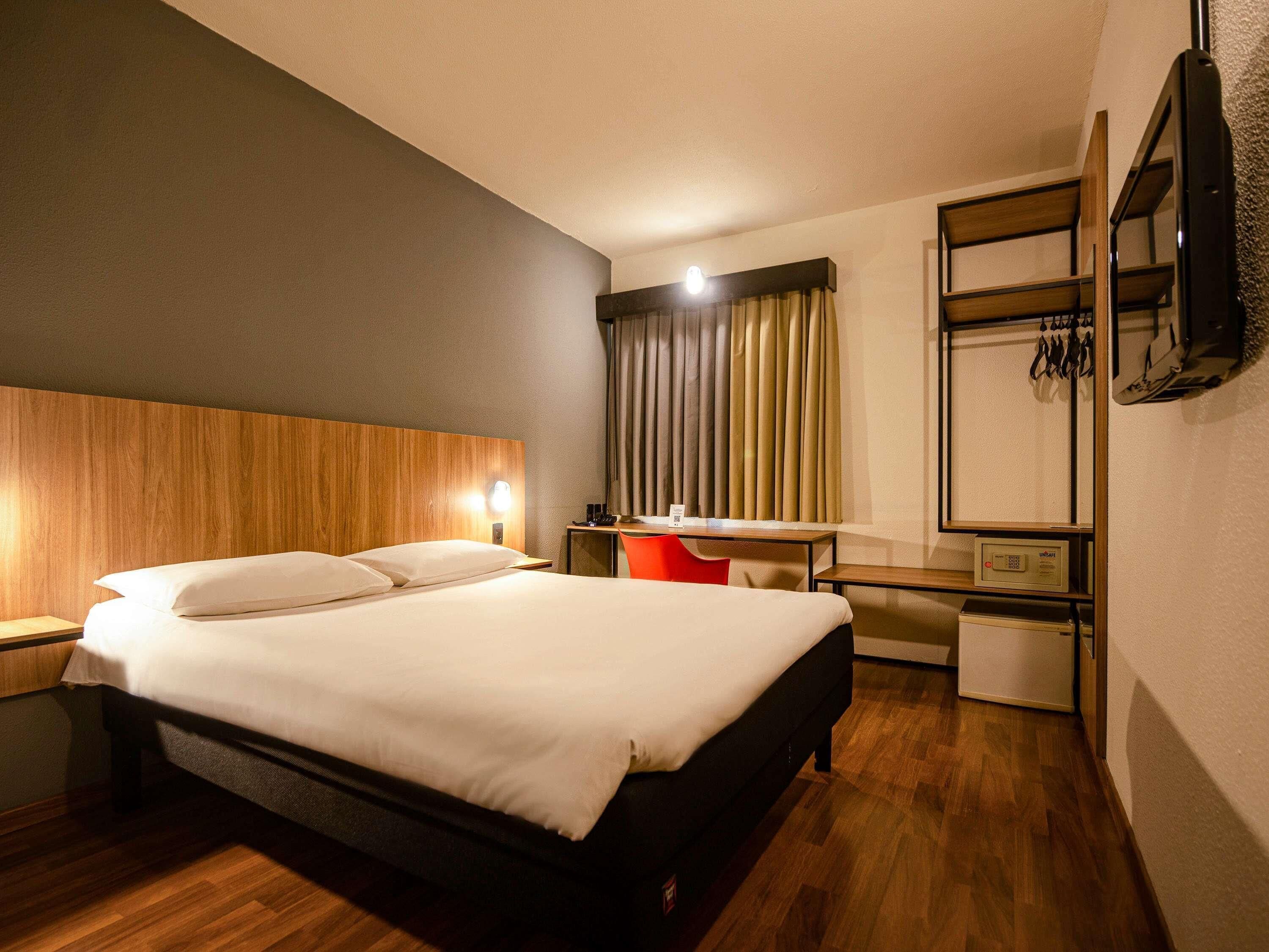 A hotel room with a bed, bedside table, lamp, desk, chair, and a TV mounted on the wall.