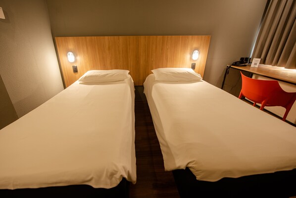 A hotel room with two single beds, a wooden headboard, a desk with a chair, and a wall-mounted phone. - ibis Betim (Betim)