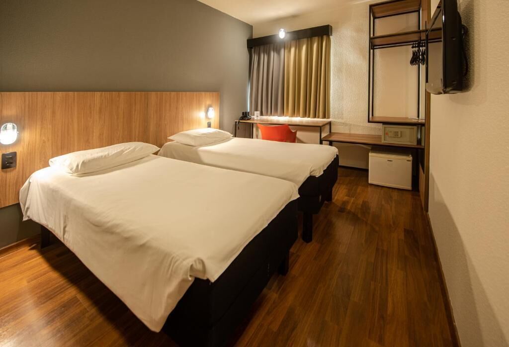 A hotel room with two beds, a desk, a chair, a small refrigerator, and a television mounted on the wall.