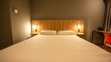 A hotel room with a large bed, two bedside lamps, a wooden headboard, and a small desk with a chair.