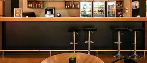 Una zona de bar con mostrador de madera, taburetes, una mesa redonda y estantes con botellas.