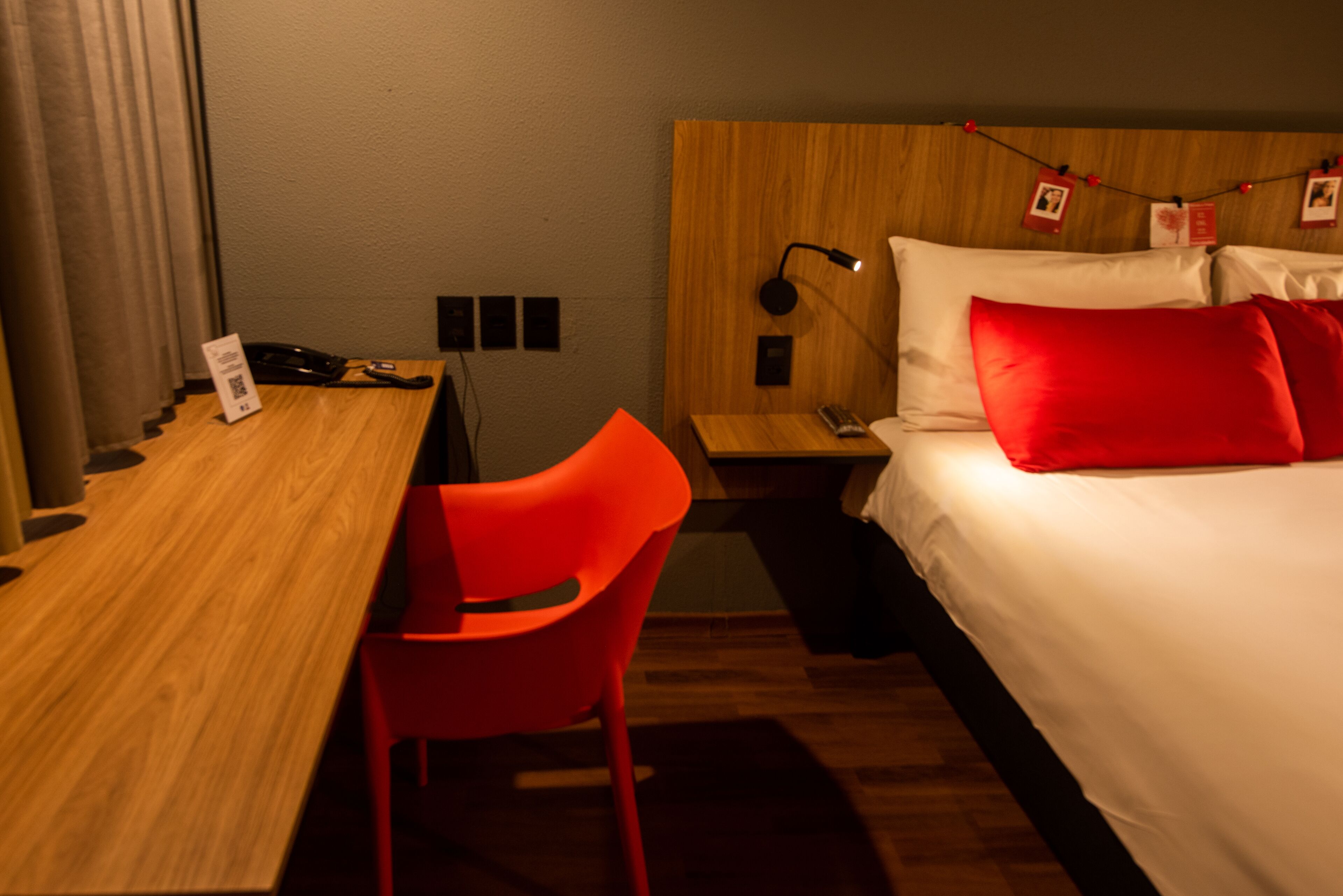 Una habitación de hotel con un escritorio de madera, una silla roja, una cama con ropa de cama blanca y roja, y una mesita de noche con una lámpara.