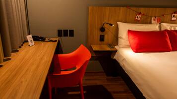 Una habitación de hotel con un escritorio de madera, una silla roja, una cama con ropa de cama blanca y roja, y una mesita de noche con una lámpara.