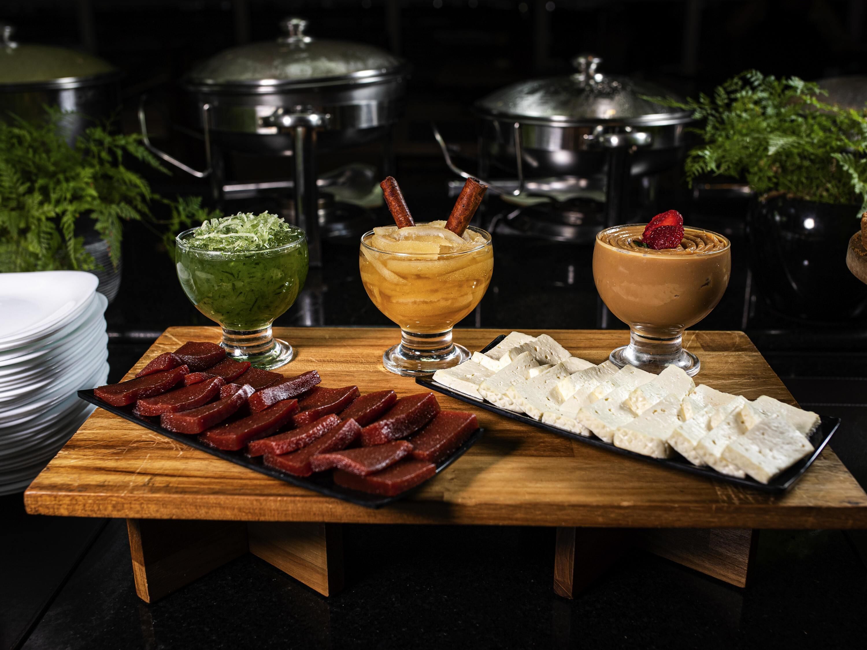 Uma mesa de madeira com três tigelas de vidro contendo bebidas de cores diferentes, um prato de carne fatiada e um prato de queijo.
