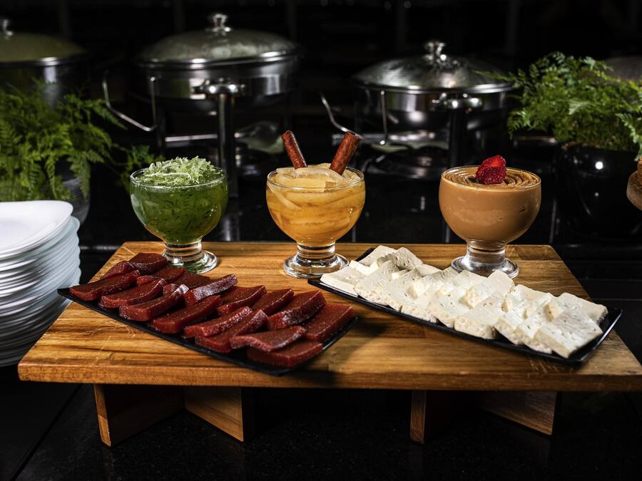 Uma mesa de madeira com três tigelas de vidro contendo bebidas de cores diferentes, um prato de carne fatiada e um prato de queijo.