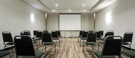 Un salón de conferencias con filas de sillas, una pantalla de proyector y piso de madera.