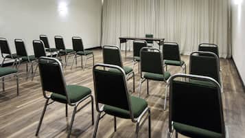 Un salón de conferencias con hileras de sillas verdes, una mesa y un telón de fondo.