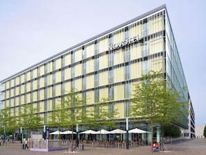 A modern hotel with a glass facade, outdoor seating area, and a clear sky. - Novotel Muenchen Messe (Munich)