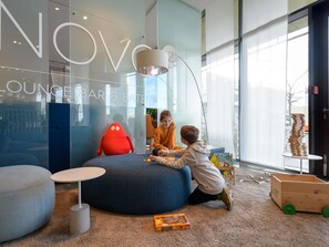 Espaço infantil com uma grande cadeira em forma de feijão vermelha, uma área de estar redonda azul e um carrinho de brinquedo de madeira.
