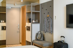 Habitación de hotel con cama, zona de estar con banco, armario, televisión y perchero en la pared.