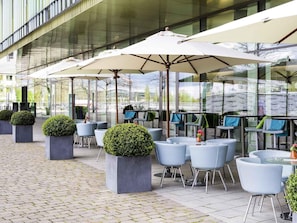 An outdoor cafe area with modern seating and large umbrellas, surrounded by glass walls. - Novotel Muenchen Messe (Munich)