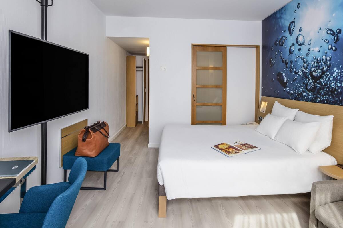 A hotel room with a bed, a TV, a blue chair, a wooden door, and a painting on the wall.