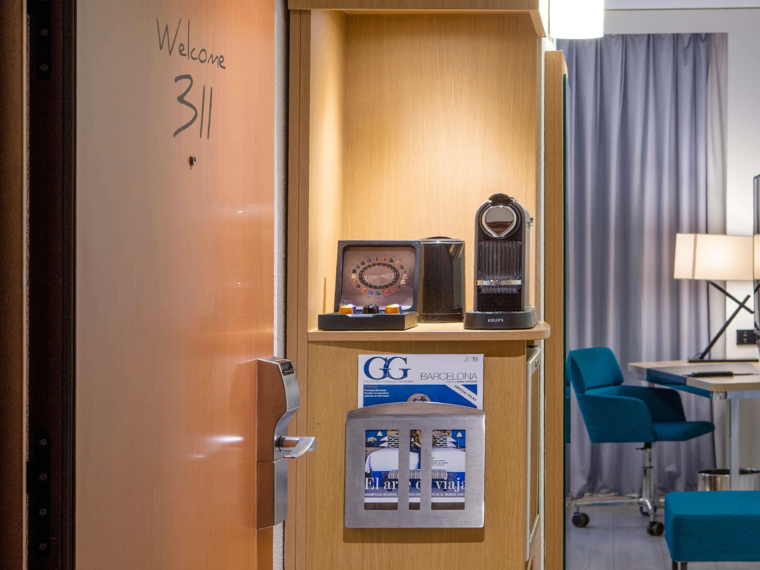 Un espace d’accueil d’hôtel avec un comptoir de réception en bois, une horloge, une machine à café Krups et un panneau indiquant la direction de Barcelone.