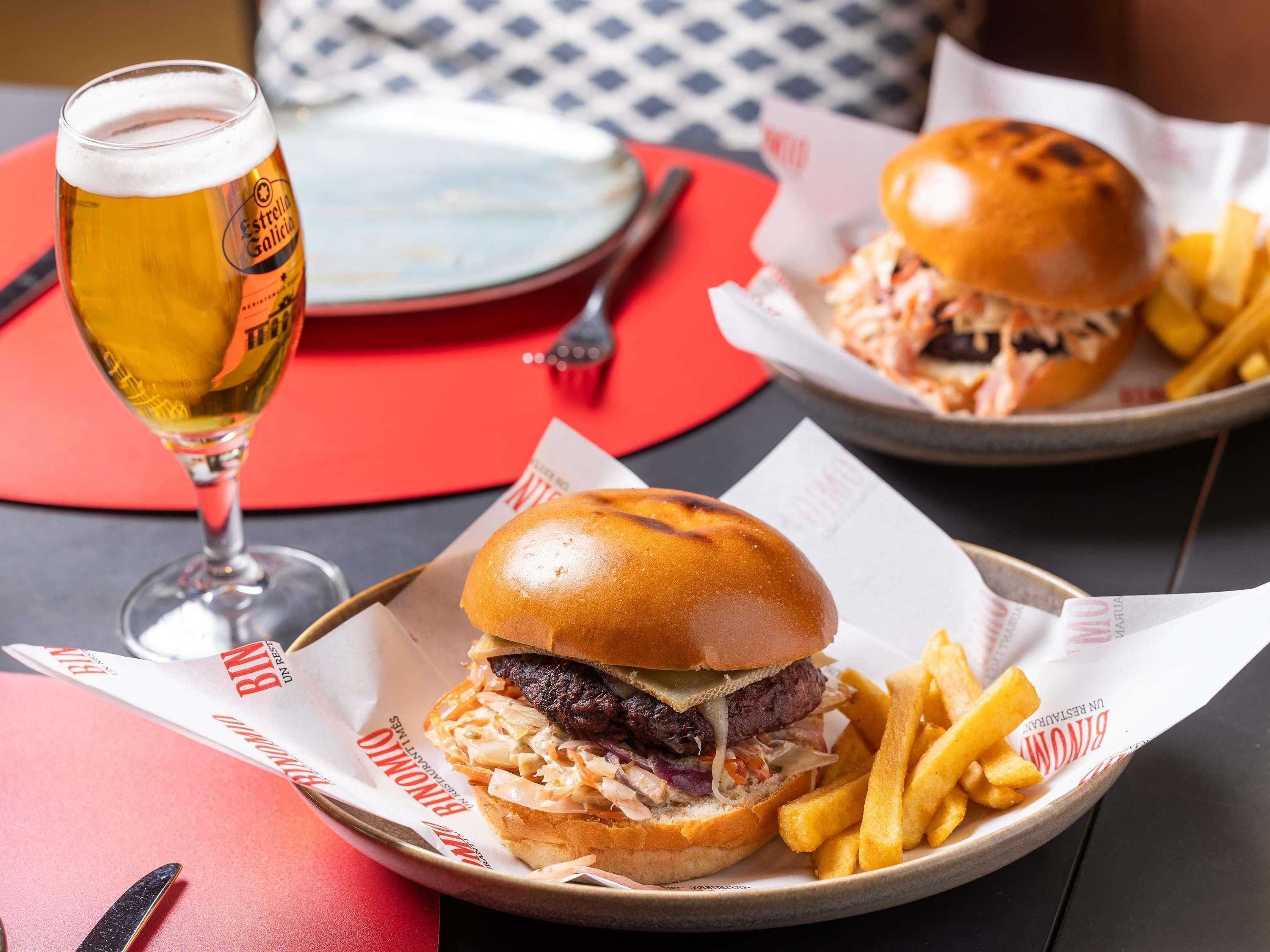 Comida que incluye hamburguesas, papas fritas y una cerveza en un vaso, sobre una mesa con un mantel rojo.