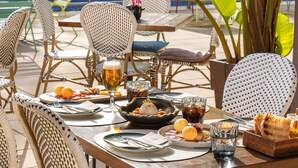 Un área de comedor al aire libre con sillas y mesas de mimbre, una piscina al fondo y diversos platos y bebidas sobre las mesas.