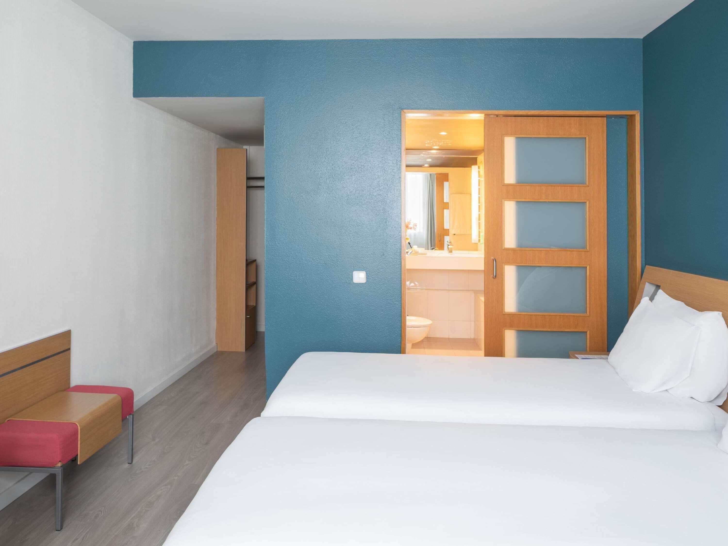 Habitación de hotel con cama, banco de madera, baño visible a través de una puerta abierta y un interruptor de luz en la pared.