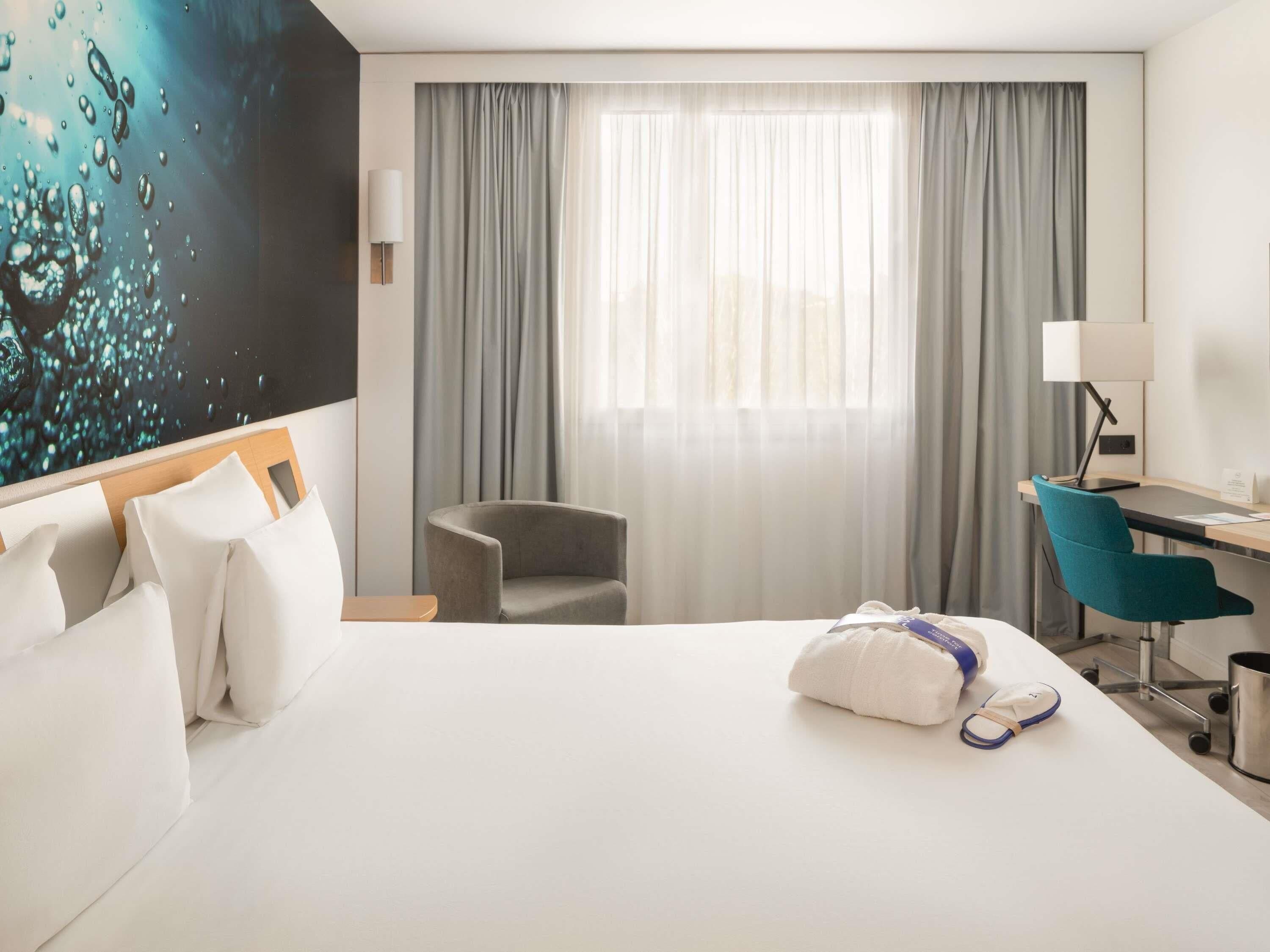 A hotel room with a bed, a desk, a chair, a lamp, and a painting on the wall.